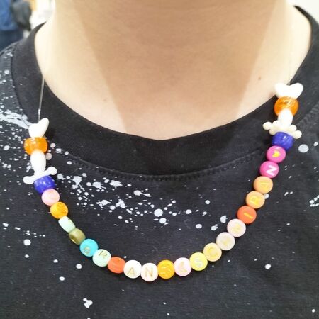 Eine Person mit einem schwarzen, gesprenkelten Shirt trägt eine bunte Perlenkette mit Buchstaben