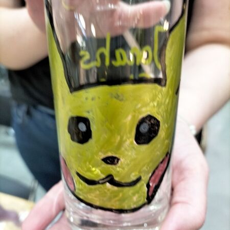 Eine Person hält ein Glas mit einem handgemalten gelben Pikachu und freundlichem Gesicht