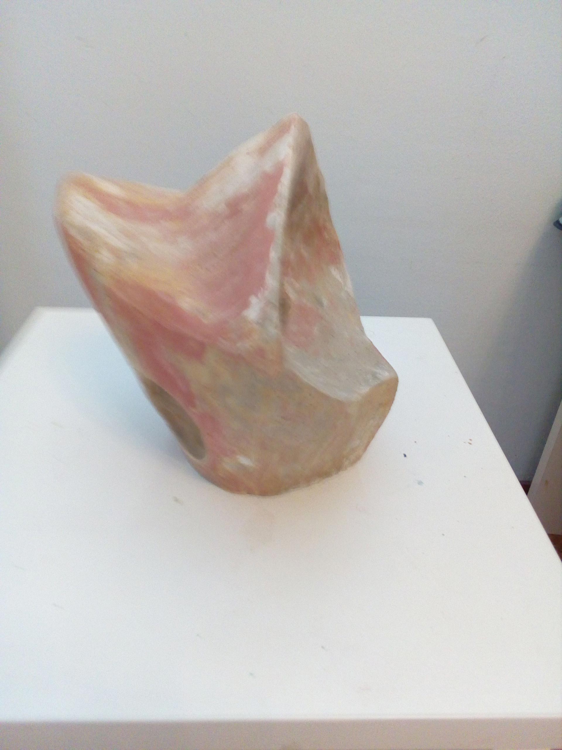 Eine abstrakte, rosafarbene Steinskulptur mit weichen Kanten auf weißem Sockel