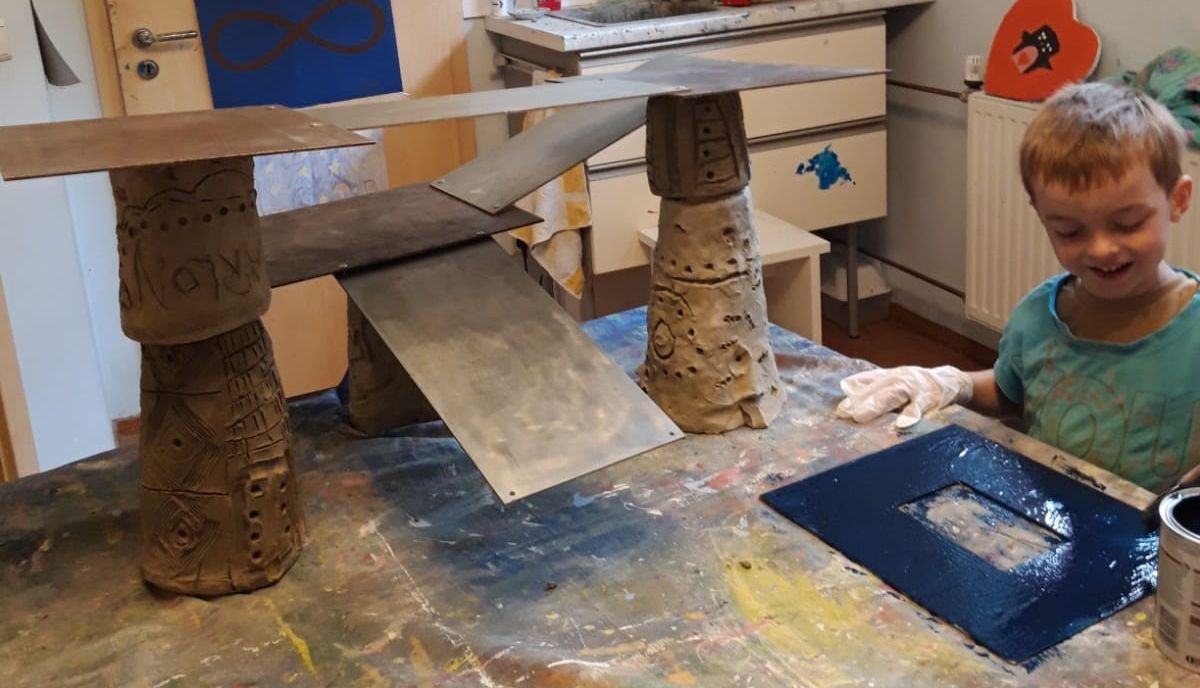 Ein Junge mit fröhlichem Gesichtsausdruck malt an einem Tisch neben einer Ton-Skulptur in Beige und Metallfarben