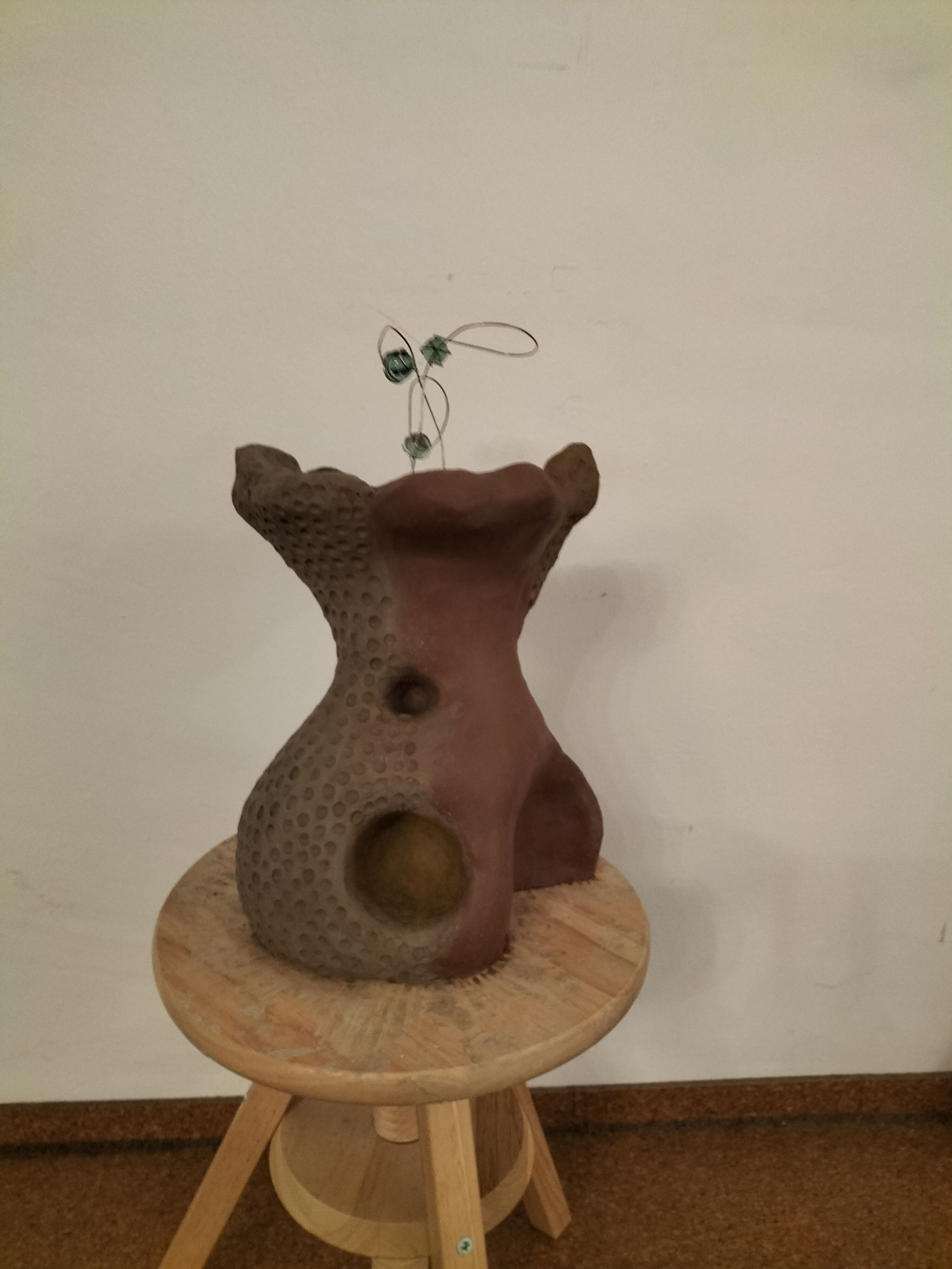 Skulptur aus braunem Ton mit runden Öffnungen und Draht auf einem Holzschemel