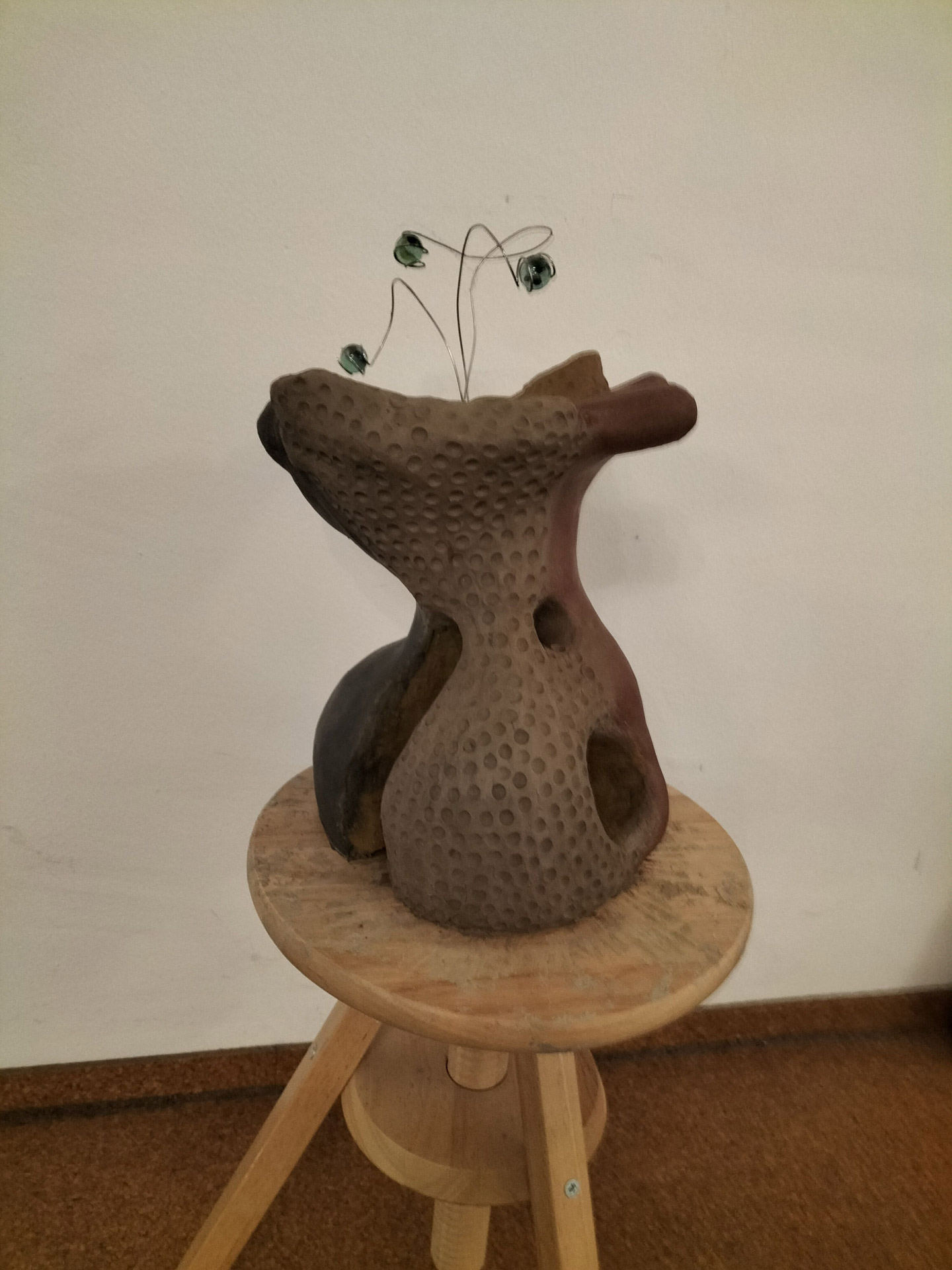 Eine handgefertigte abstrakte Skulptur mit löchriger Oberfläche und Drahtblumen auf hellem Hocker