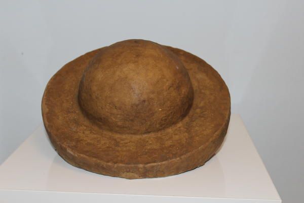 Ein brauner, halbrunder Hut mit breiter Krempe aus festem Material auf weißem Podest