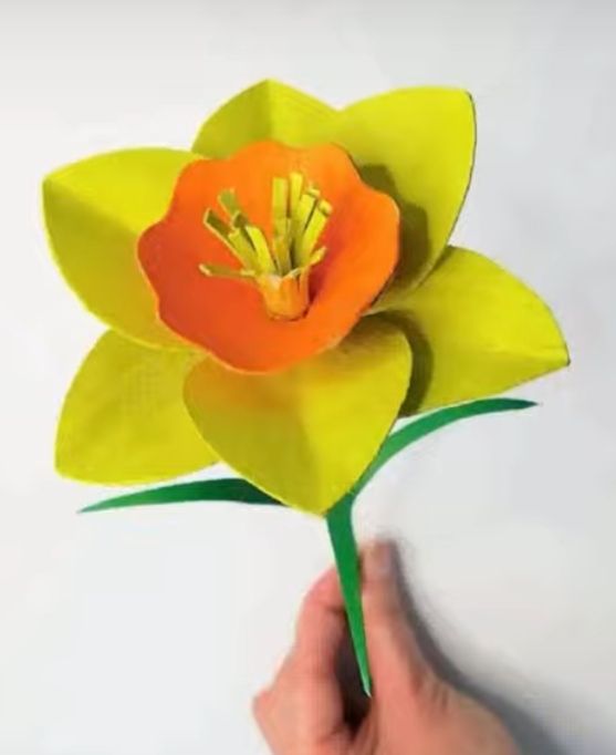 Hand haelt gelbe Papierblume mit orangefarbener Mitte und Stiel