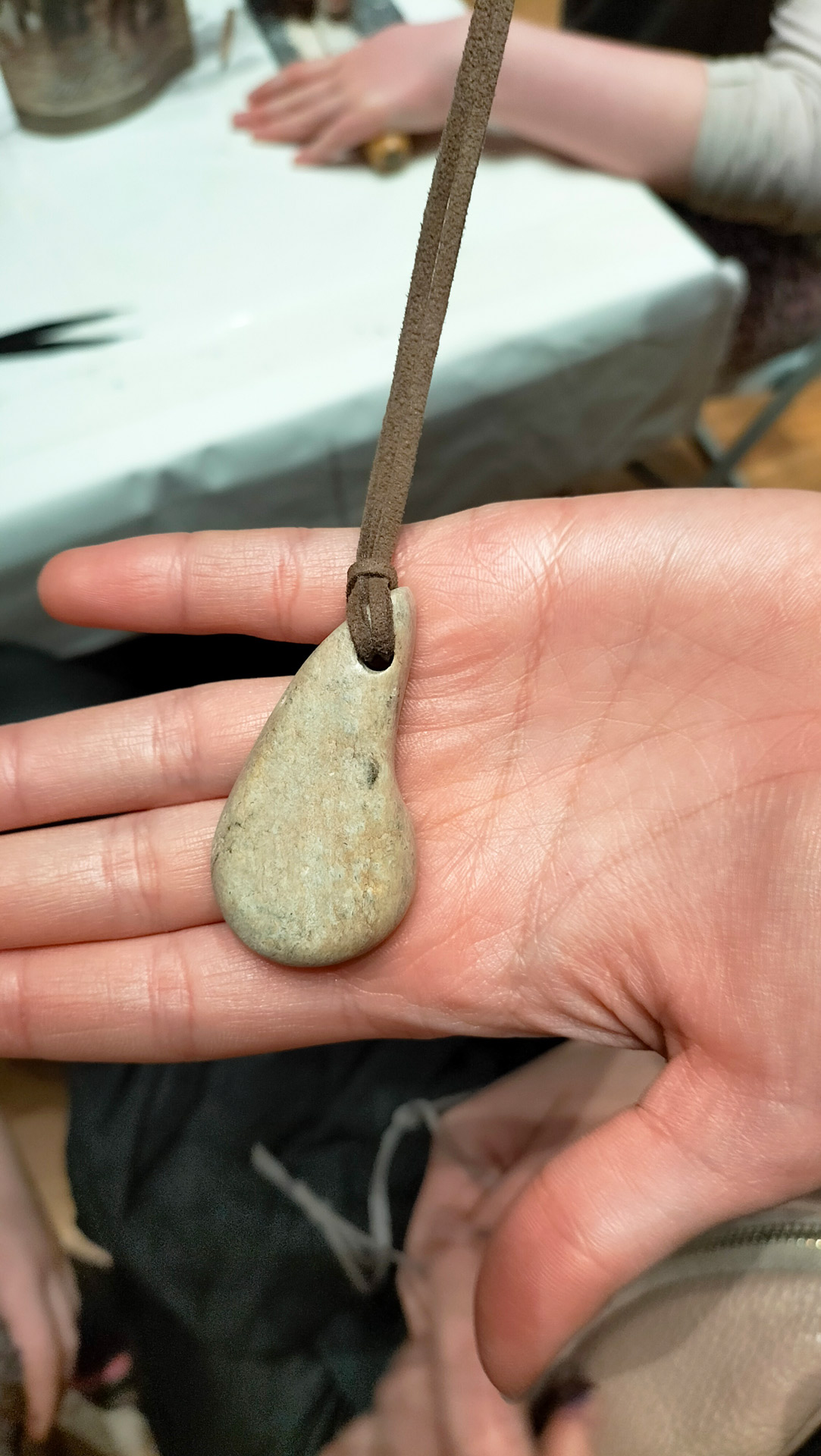 Hand mit neutralem Ausdruck haelt grauen Tropfenanhaenger am Lederband
