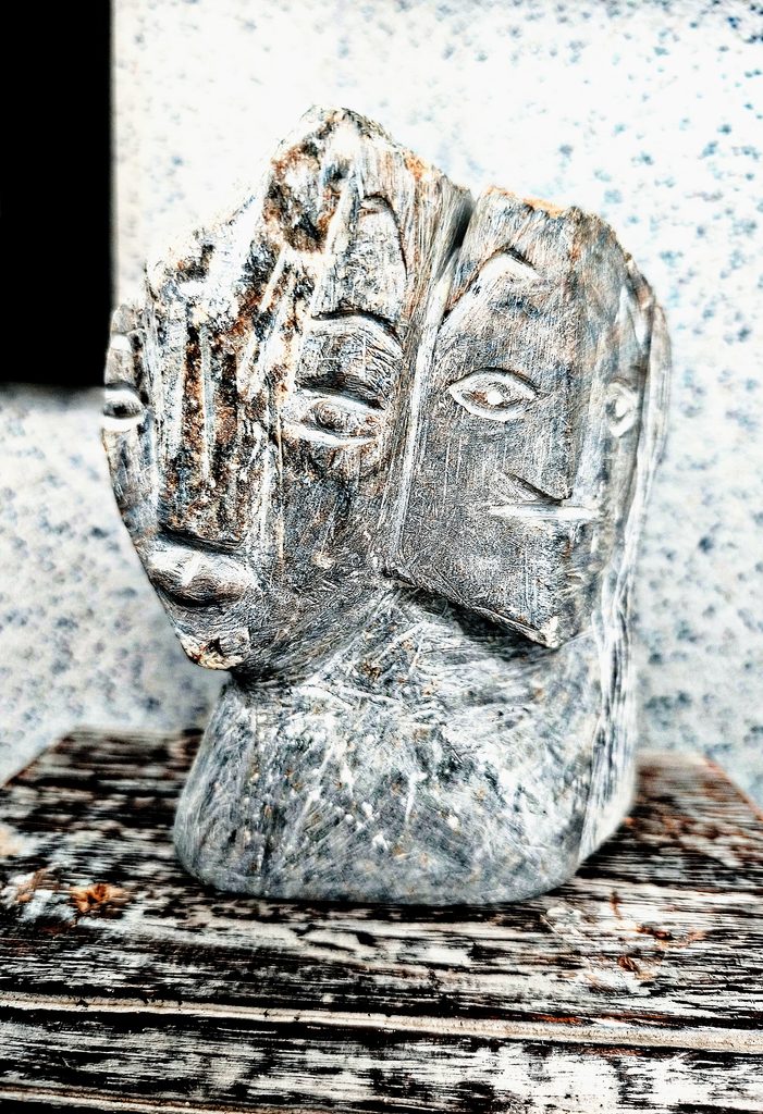 Eine graue Steinskulptur mit zwei geschnitzten Gesichtern auf Holzuntergrund
